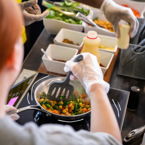 A person in disposable gloves stirs chickpeas, spinach, and carrots in a skillet on an induction burner, with bowls of fresh ingredients lined up nearby.
