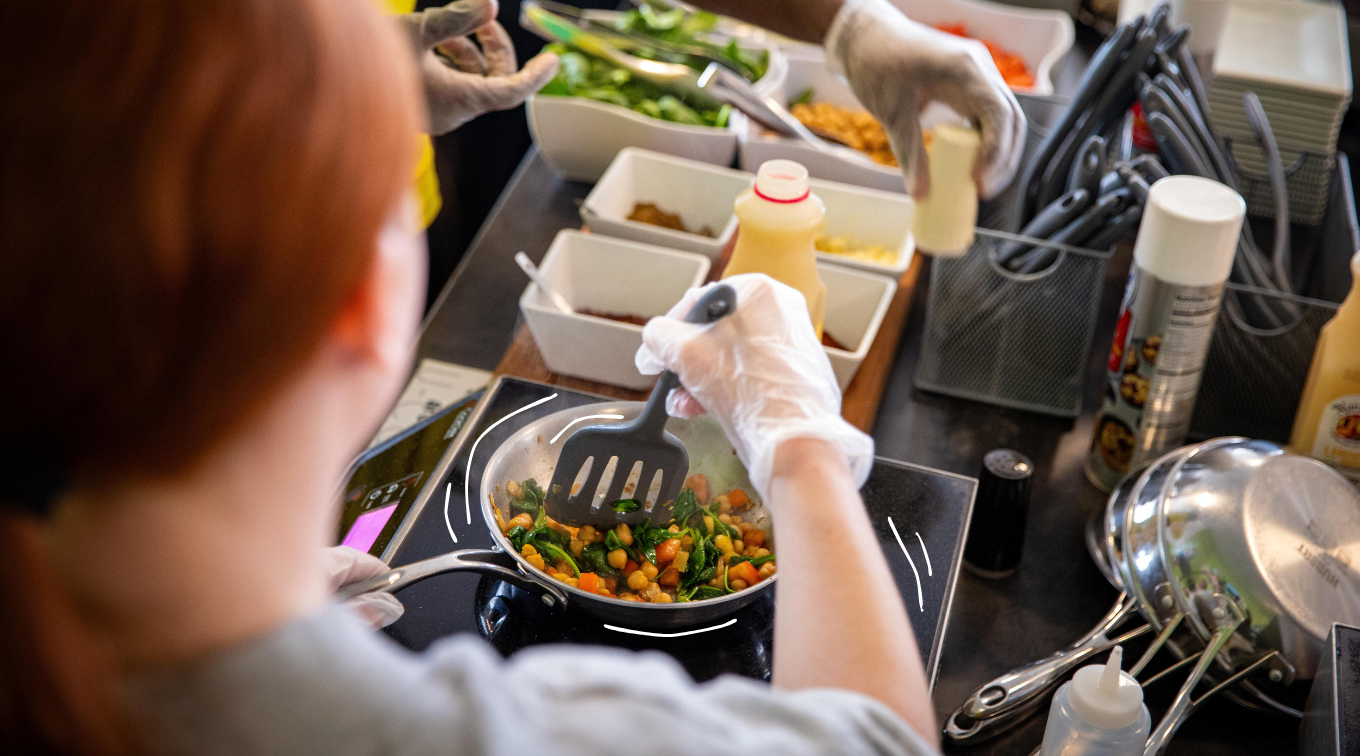 A person in disposable gloves stirs chickpeas, spinach, and carrots in a skillet on an induction burner, with bowls of fresh ingredients lined up nearby.
