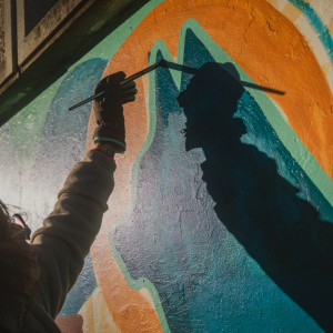 An artist in a work glove reaches up with a paintbrush to add detail to the mural, their shadow stretched across the wall in low sunlight.
