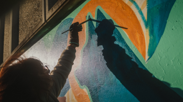 An artist in a work glove reaches up with a paintbrush to add detail to the mural, their shadow stretched across the wall in low sunlight.