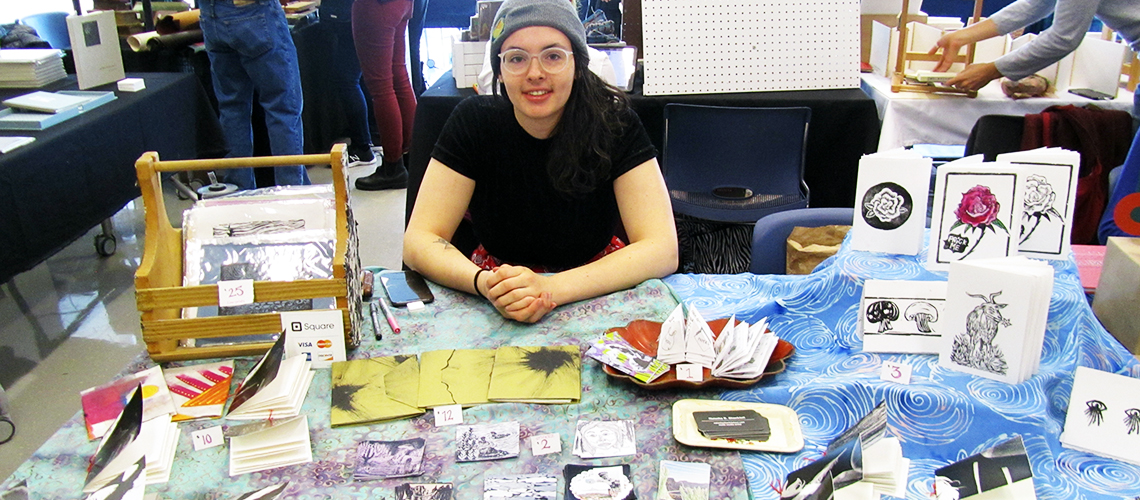 Image of a Book Arts Bazaar exhibitor sitting at a table featuring their artist books, zines, and paper objects.