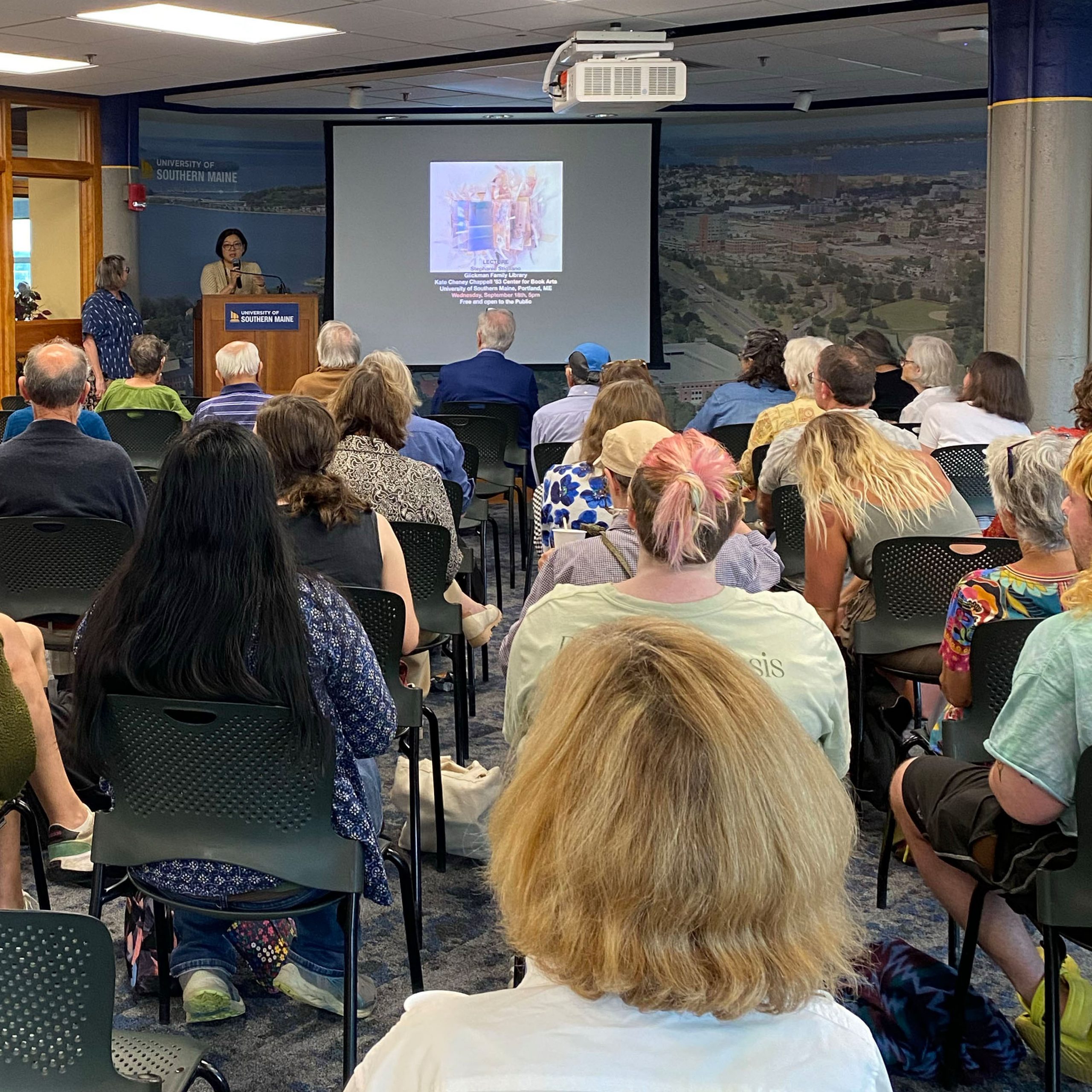 Photograph of an artist talk with Stephanie Stigliano featuring a room full of attendees and a projection screen displaying samples of Stigliano's book arts work.