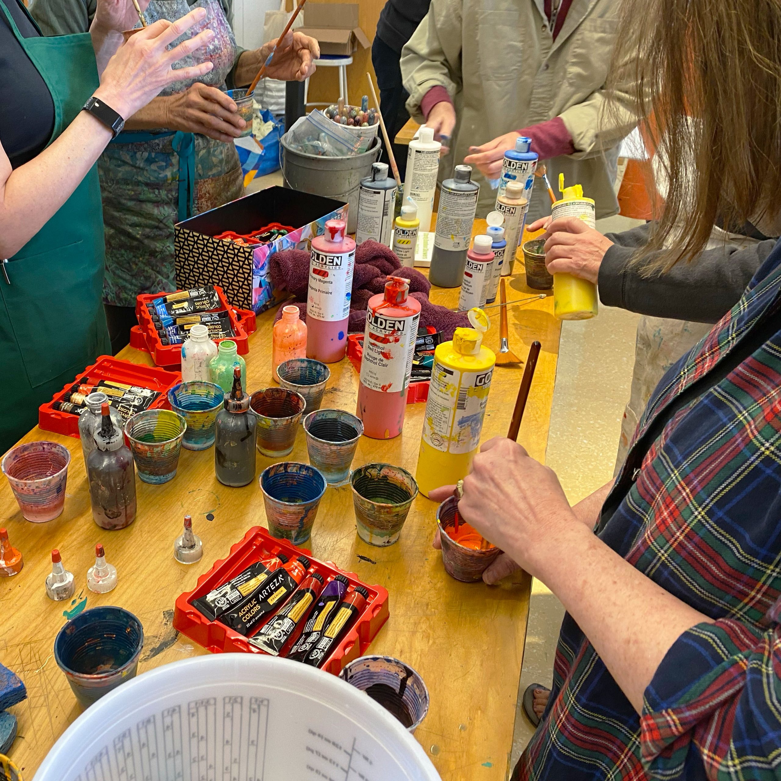 Photograph of a workshop in-session showing hands mixing paint around a table crowded with paints, brushes, and cups of pigment.