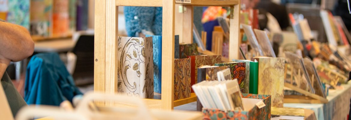 A vendors display table viewed from a side perspective. The table features handmade books of all colors and sizes, with some handmade paper displayed alongside the tiers of items.