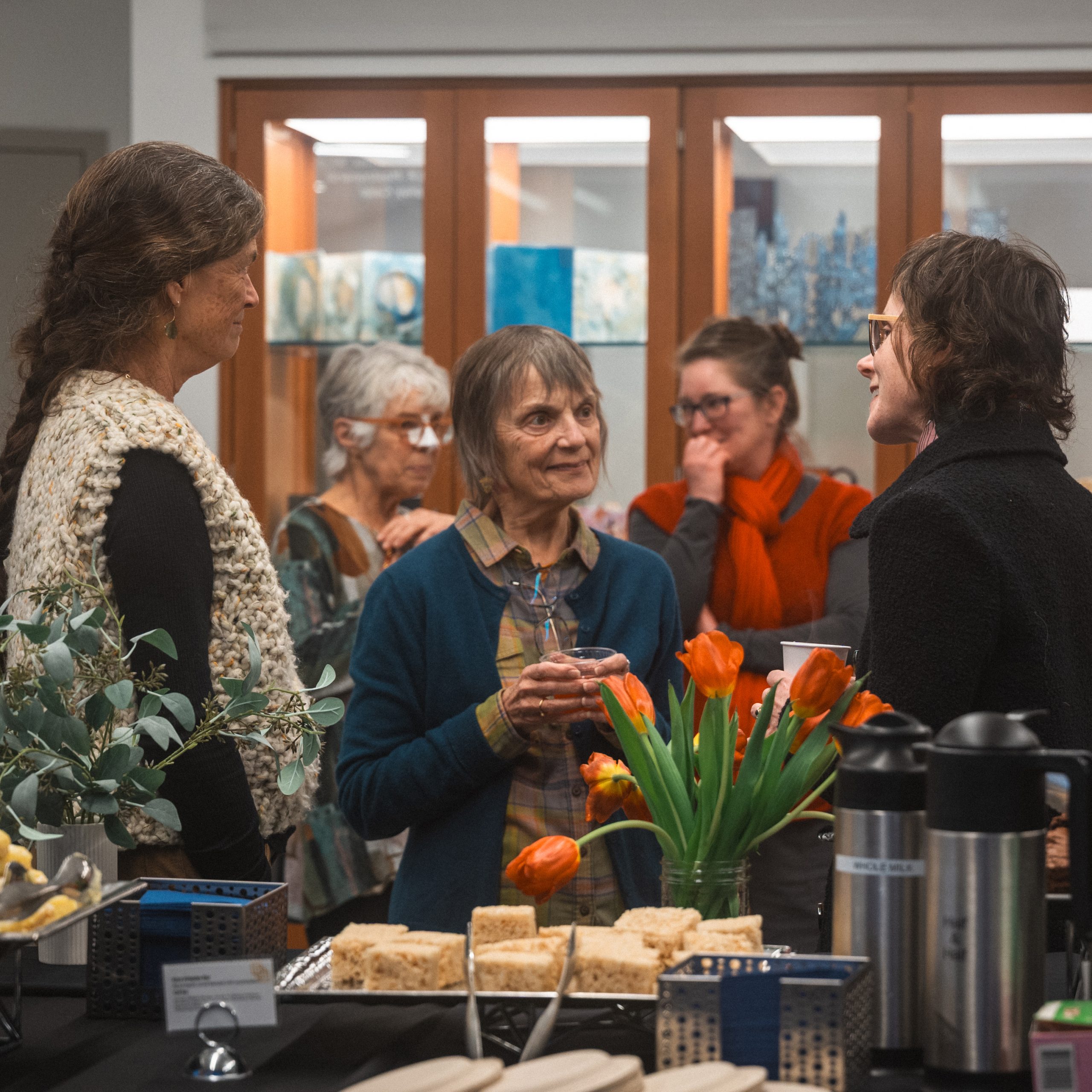 Three people stand in the foreground, with two in the back. All people seem to be actively in conversation, behind a table of snacks in the Kate Cheney Chappell Center for Book Arts Studio.