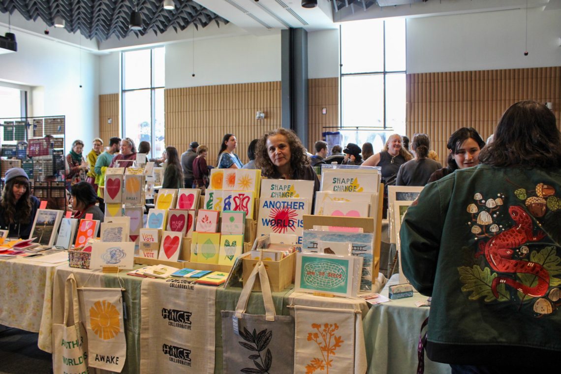 Elizabeth Jabar from Hinge Collaborative stands behind her table (covered in colorful, handmade prints) in a crowded event space at the 2026 Book Arts Bazaar