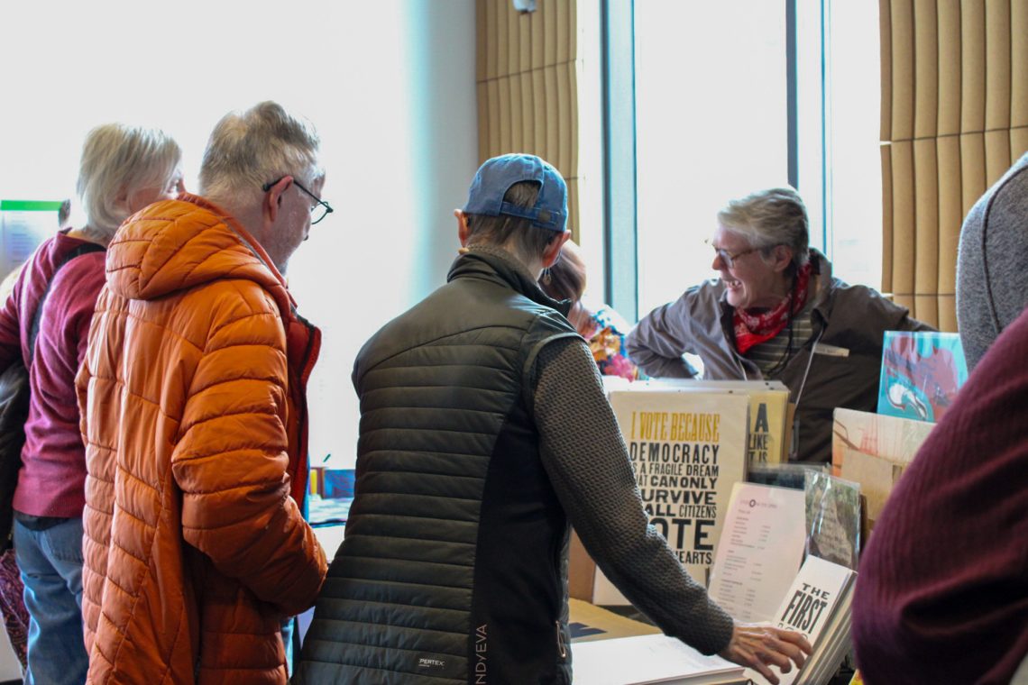 Cynthia Marsh stands behind her table displaying letterpress prints while talking with event attendees.