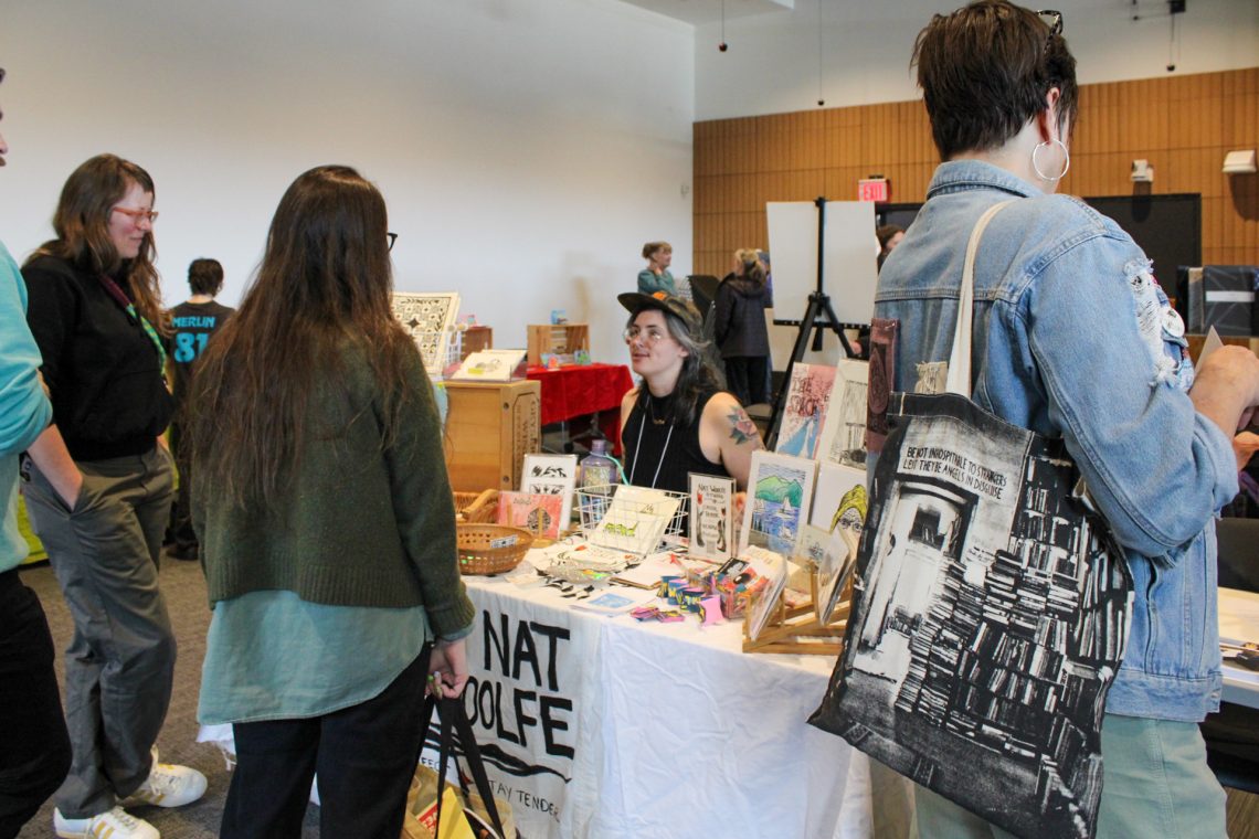 Nat Woolfe sits behind their vendors table with handmade prints and stickers while talking to event attendees.