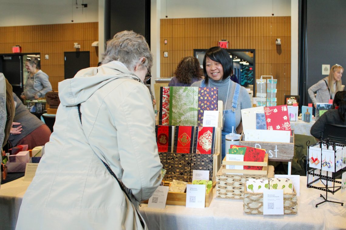 Evelyn Wong stands behind their vendors table displaying handmade books of all sizes. An event attendee examines the table closely.