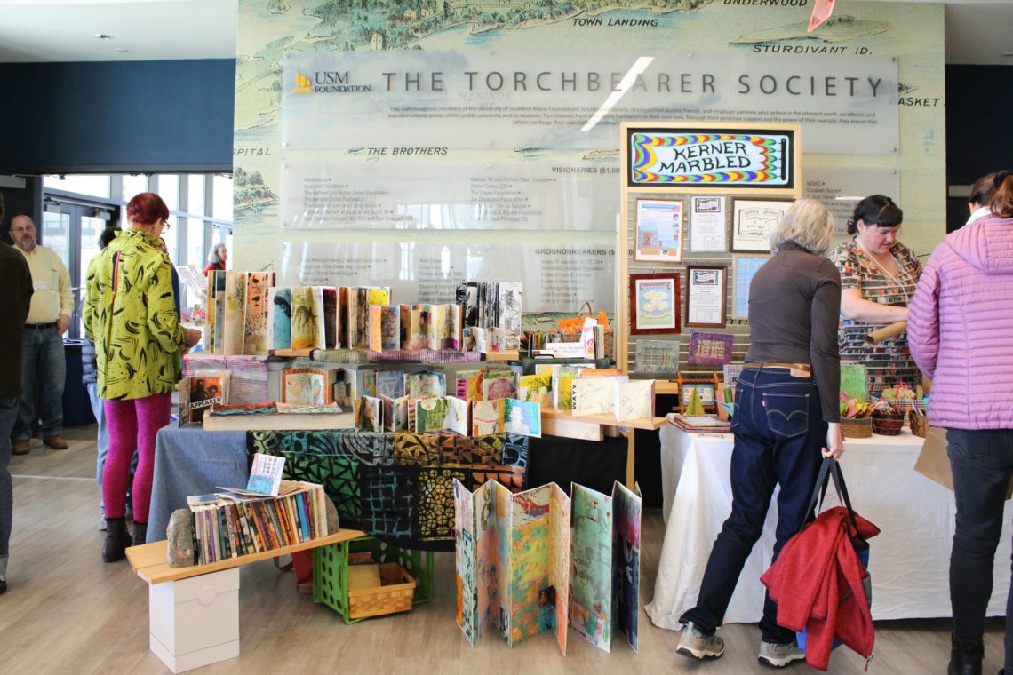 Meredith Mustard and Kerner Marbled exhibit side-by-side at the 2026 Book Arts Bazaar, exhibiting colorful artist books and hand-marbled paper. Event guests stroll by.
