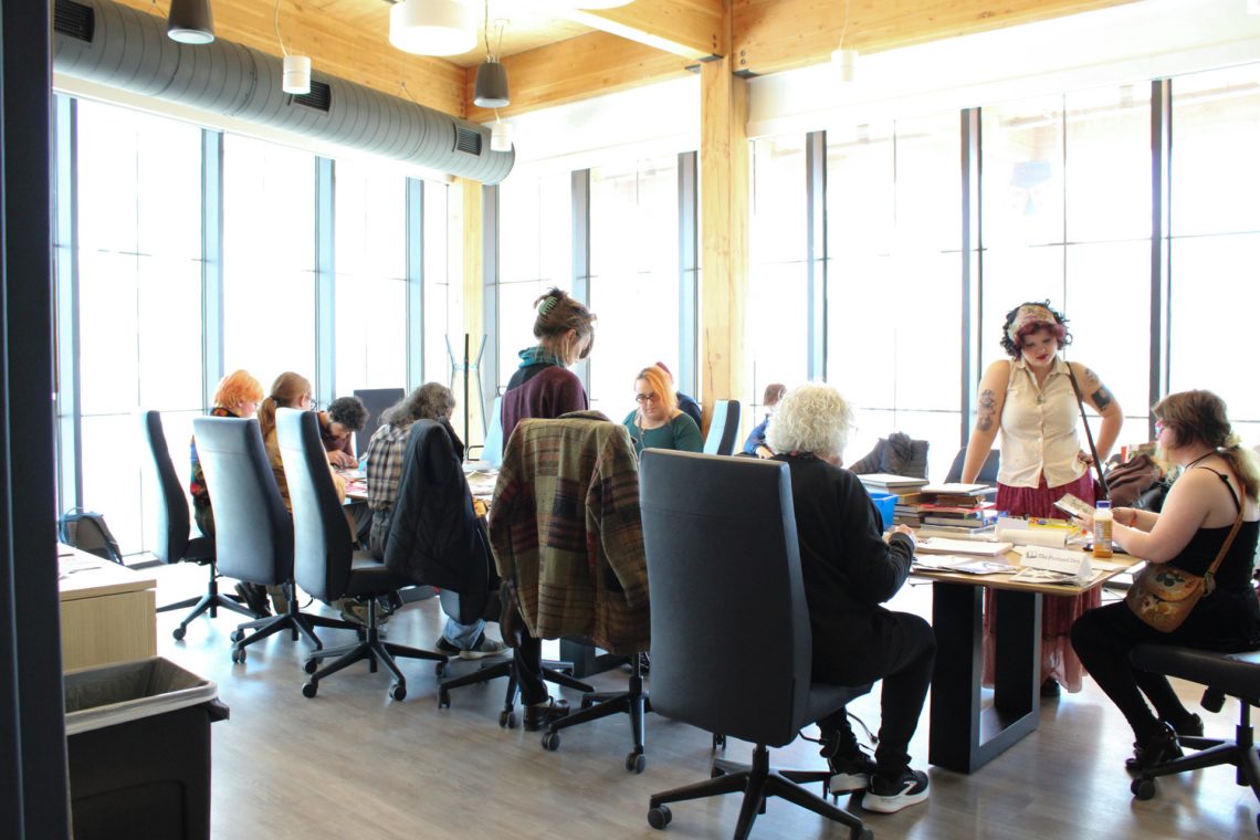 Event attendees sit in the Book Bazaar workshop room at a long table while making collages and zines while in conversation with one another.