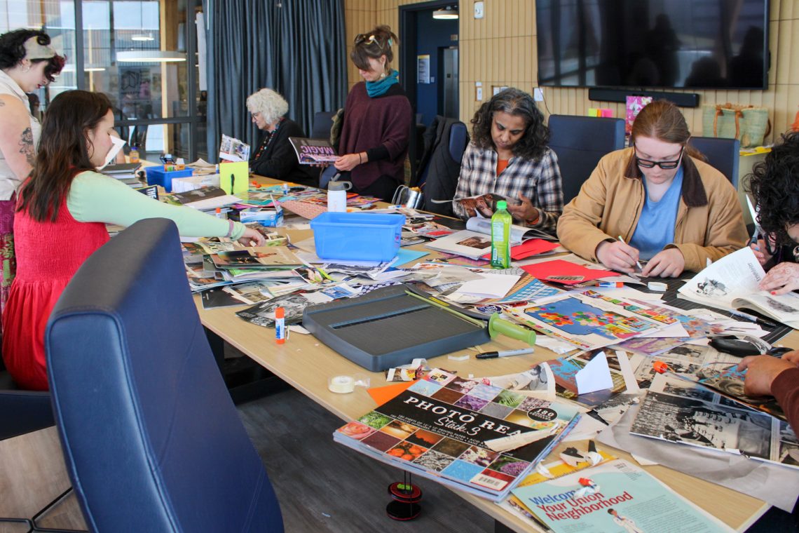 Event attendees sit in the Book Bazaar workshop room at a long table while making collages and zines while in conversation with one another. This photo takes a closer look at the workshop materials on the table, which include magazine scraps, glue sticks, pens, and colorful paper.