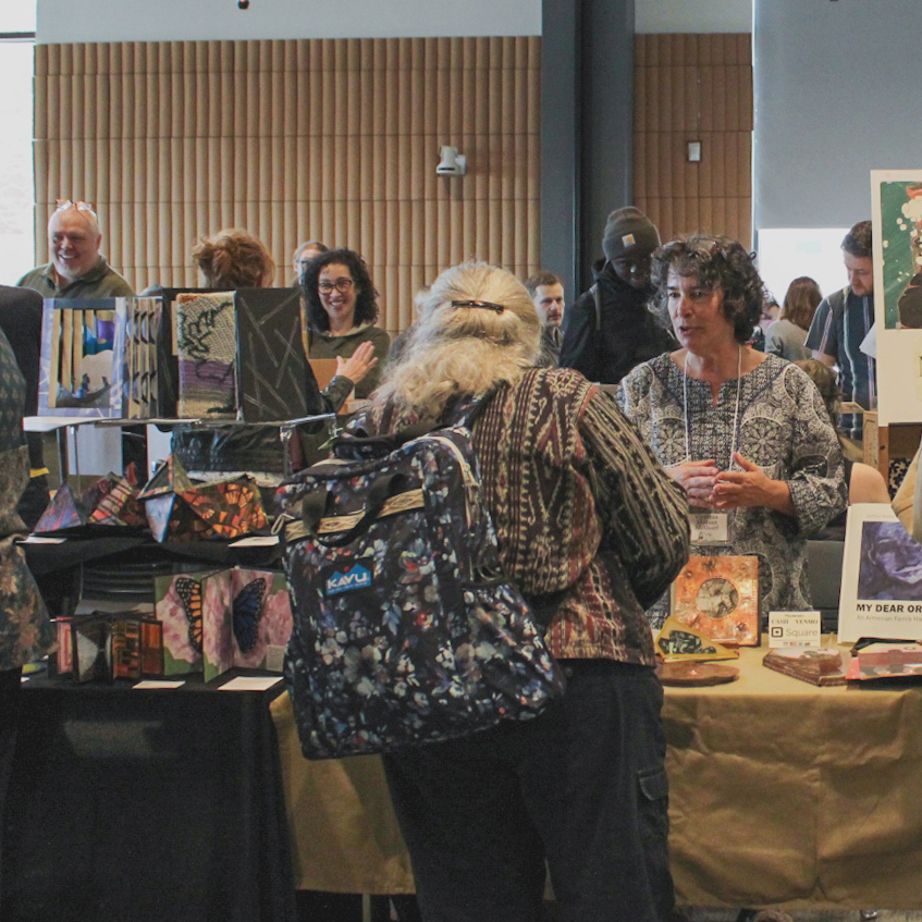 A crowded exhibition hall shows book vendors at tables surrounded by customers and student visitors. The foreground shoes a person with their back turned toward the viewer, busy in conversation with the book vendor.
