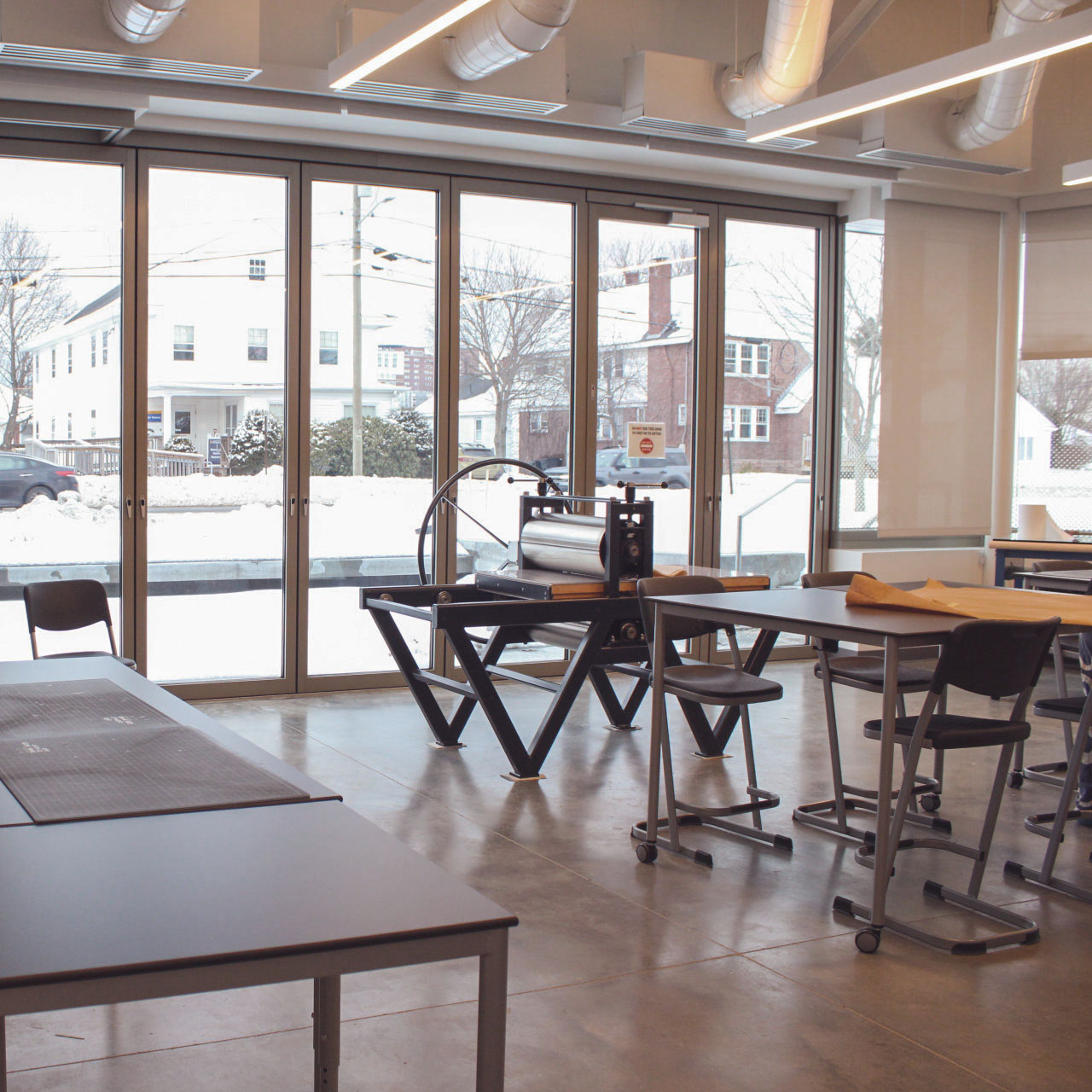 Photograph of the Kate Cheney Chappell Center for Book Arts Studio in the Crewe Center for the Arts located on the University of Southern Maine Portland campus. The studio space is empty, facing a wall of windows behind an etching press and classroom tables and chairs.