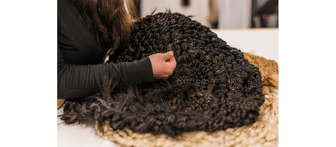 A long white table with braided artifical hair in blonde and black is laid out. An arm with a long black sleeve reaches out to continue braiding. The braids are in circles, like trivets.