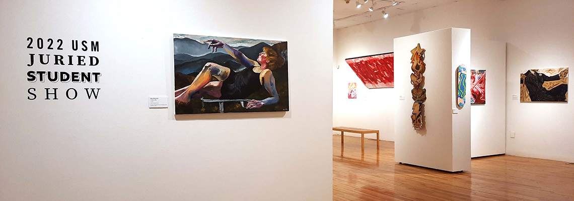 An image of a gallery with artwork on the walls. At left a sign reads "2022 JURIED STUDENT SHOW". To its right, a woman reclines.