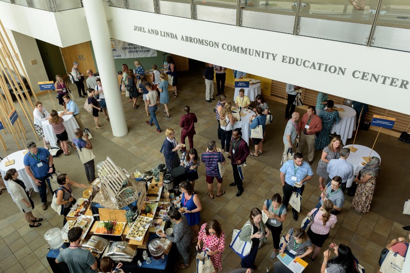 a birds-eye-view of folks mingling and eating at an event in a room labeled the Joel and Linda Abromson Community Education Center