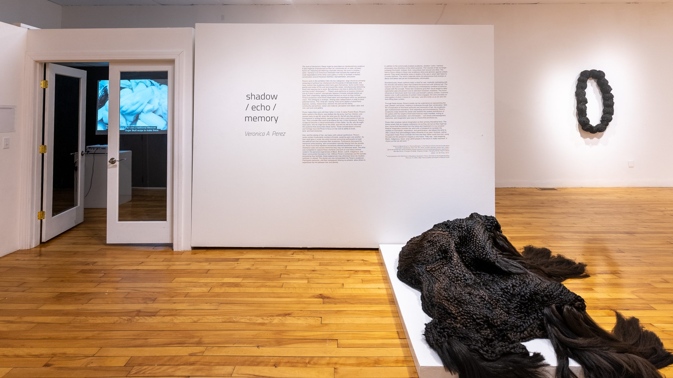 A gallery with white walls and wooden floors. At left: a projection of white clay hands, in middle: vinyl exhibition text; at right: a hair sculpture on a low plinth