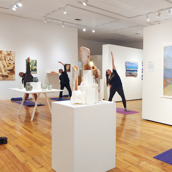 A white walled gallery with wooden floors. Three students engage in a stretching exercise on yoga matters surrounded by hanging artworks and sculptures on pedastals.