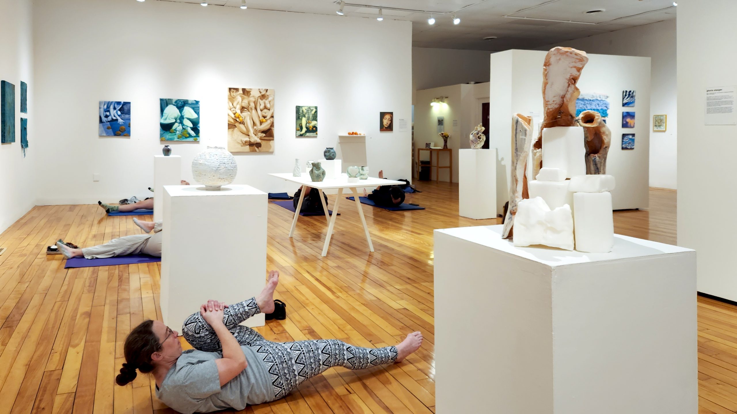 A white walled gallery with wooden floors. Attendees engage in an exercise on yoga mats surrounded by hanging artworks and sculptures on pedastals.