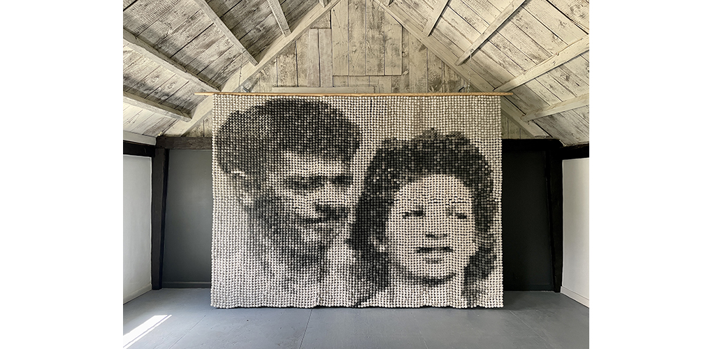 A paper beaded tapestry featuring a black and white portrait of a man and a woman's faces, standing side by side. The overall tapestry is in a barn.