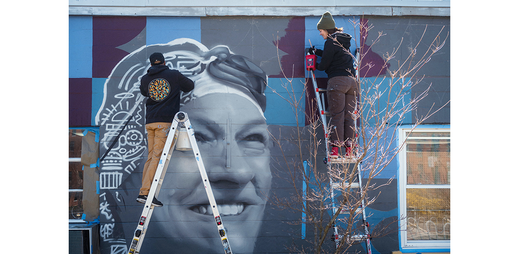 Two people on ladders paint a mural on a wall featuring a vectorized black and white portrait of a woman in a swim cap