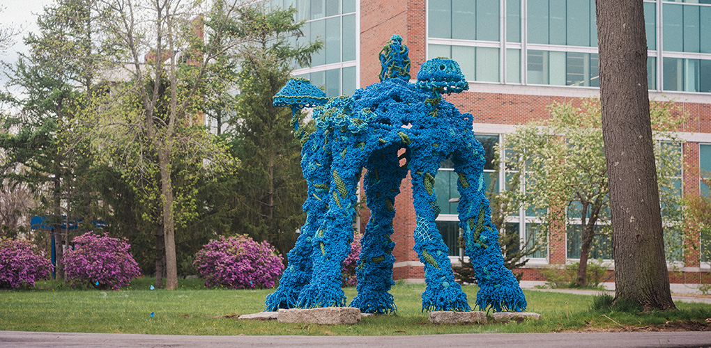 A blue and green quadriped made out of marine rope towers 14' tall over a campus green.