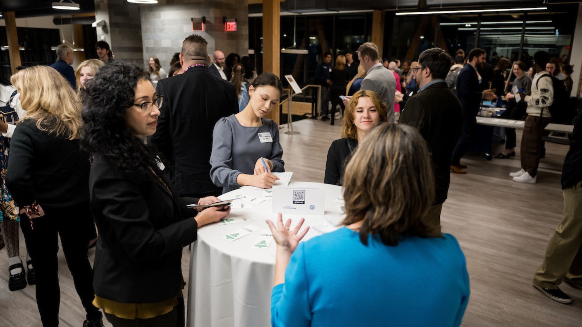 Students and employer representatives stand around a cocktail table talking, multiple other people stand around in conversation.