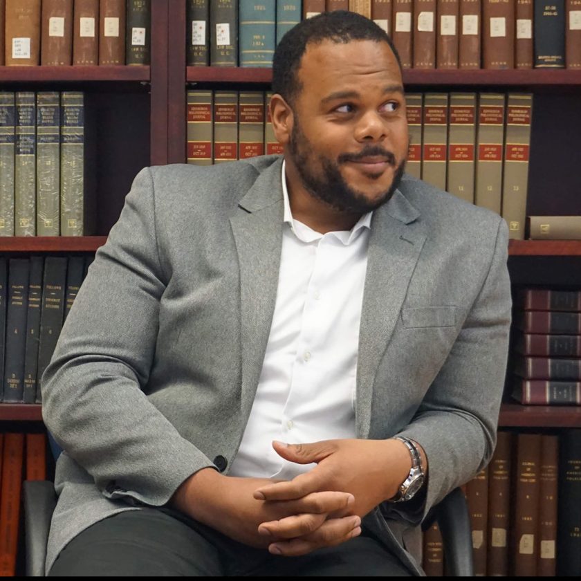 Dr. William Paris of Toronto University is seated with hands folded in front of a shelf of books.