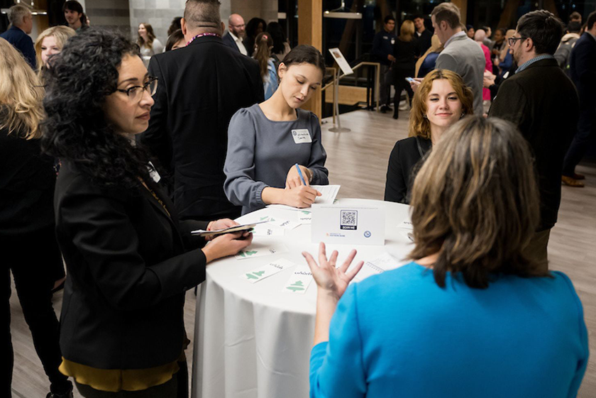 Students and employer representatives stand around a cocktail table talking, multiple other people stand around in conversation.