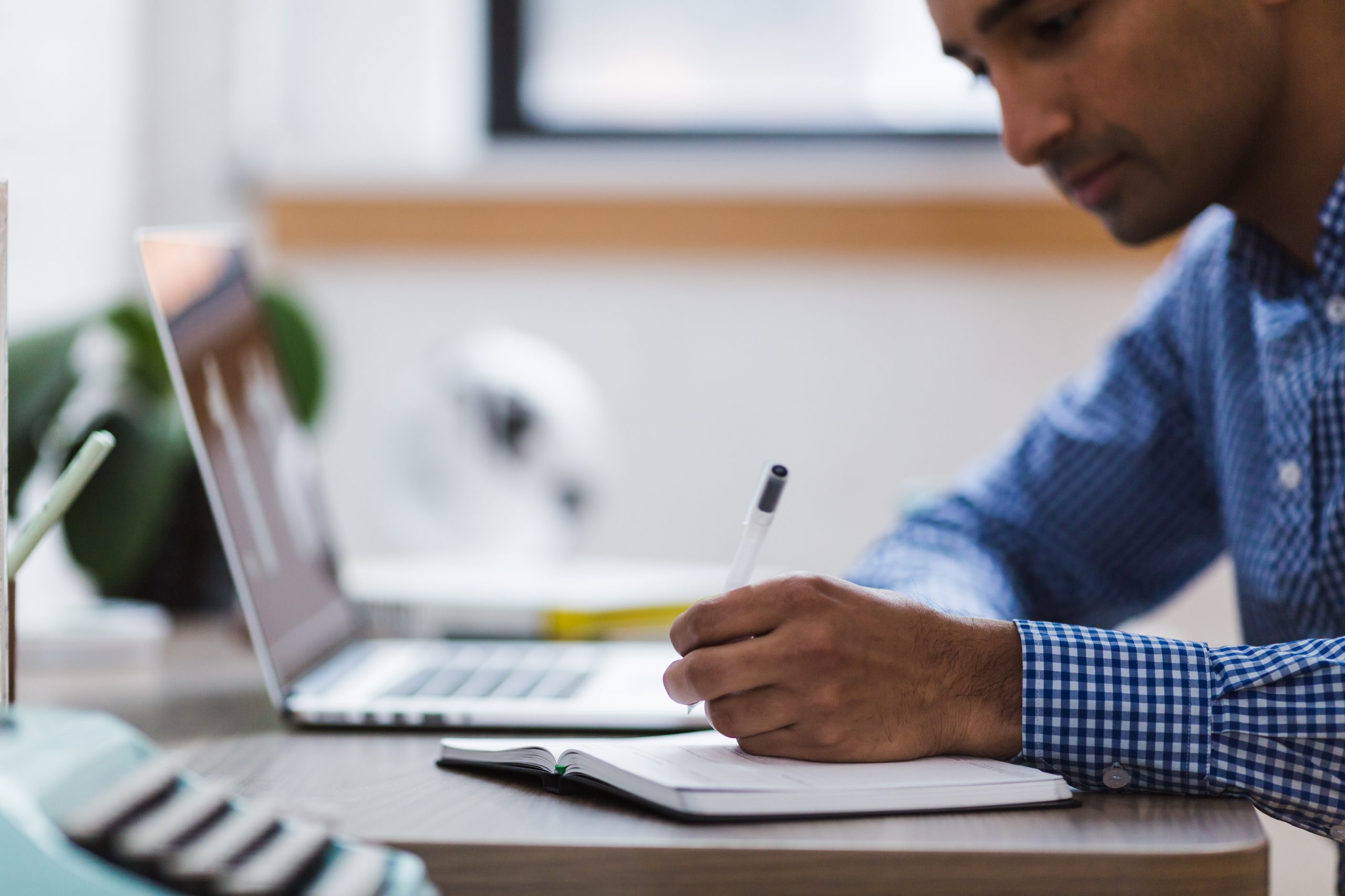 a person sits in front of an open laptop at a desk, while they hold a pen to an open book