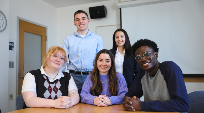 March 25, 2026 Event Photo for Future Leaders’ Voices: Student Perspectives on Pluralism and Democracy Panelists: Mia Noon, Haley Greco, Djino Kavula Nzau, Justin Gibbons, Ashley Gonzalez in a USM conference room three are sitting at a conference table and two are standing behind them.