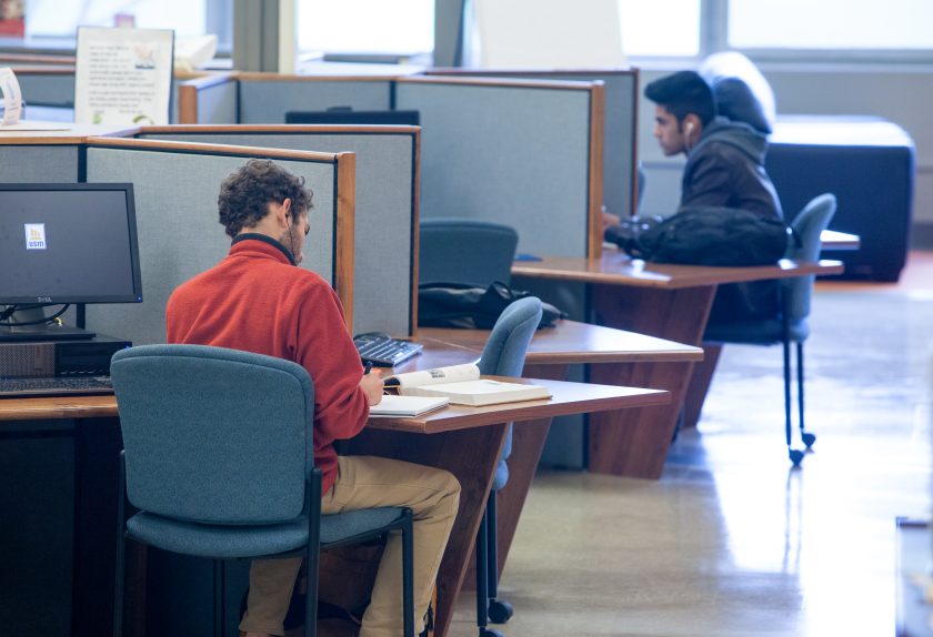 In a library setting, two students sit at separate, cubicle-like workstations