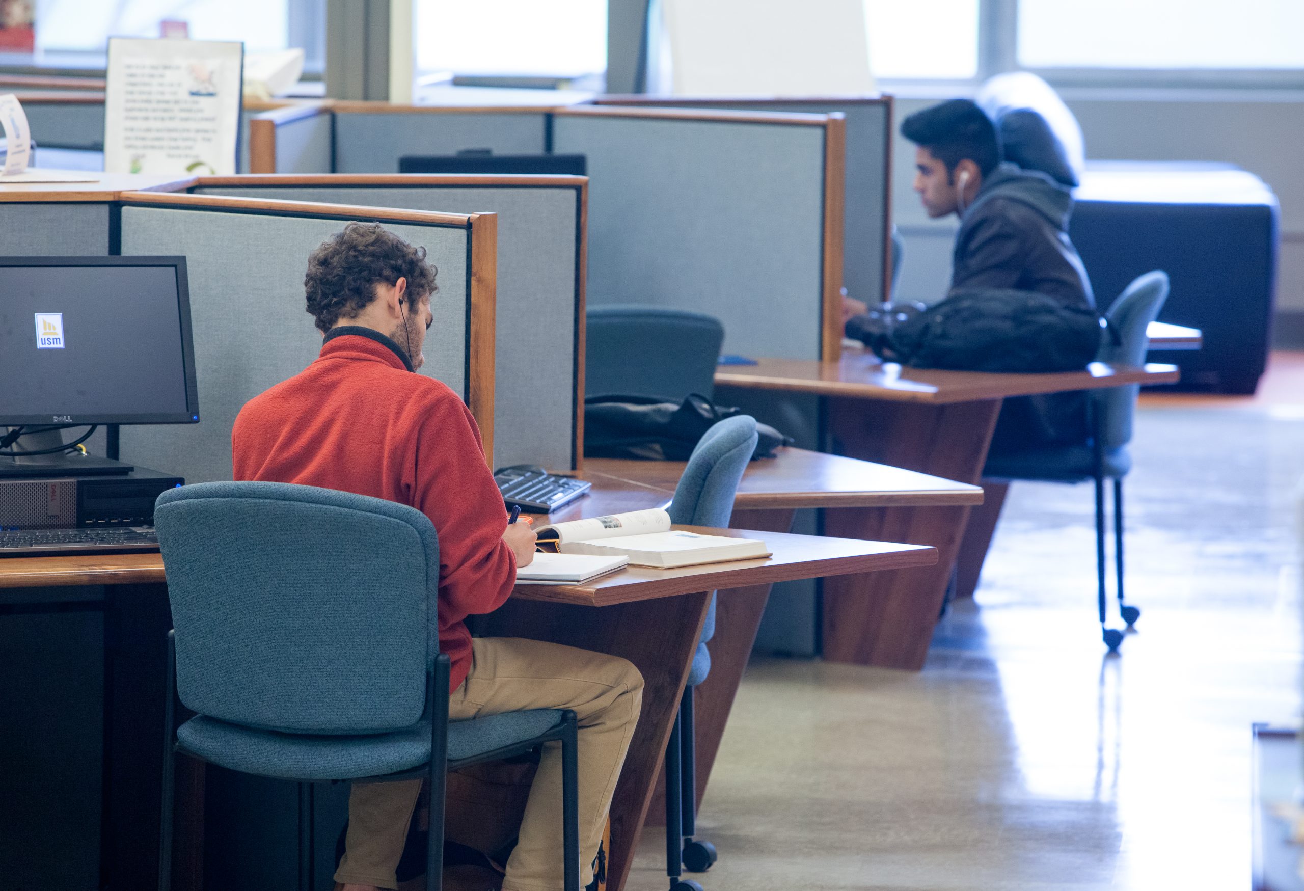 In a library setting, two students sit at separate, cubicle-like workstations