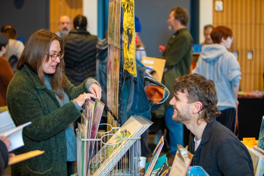 Photo from the 2025 Book Arts Bazaar of two people, one perusing art prints the other sitting at a vendors table.