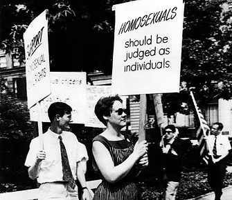 Black and white image of protestors from 1965. One sign reads, "Homosexuals should be judged as individuals."