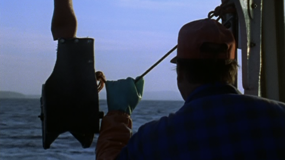 Closeup of man facing away from the camera on a fishing boat. He is silhouetted against a blue-purple sky and dark blue water.