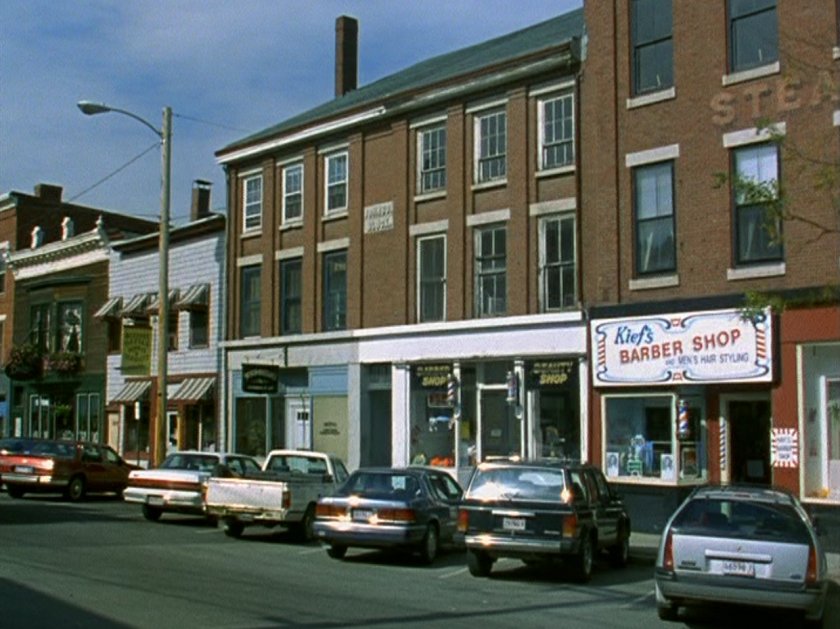 Photo of a downtown street with '90s era cars parked in front of 3 and 4-story brick buildings.