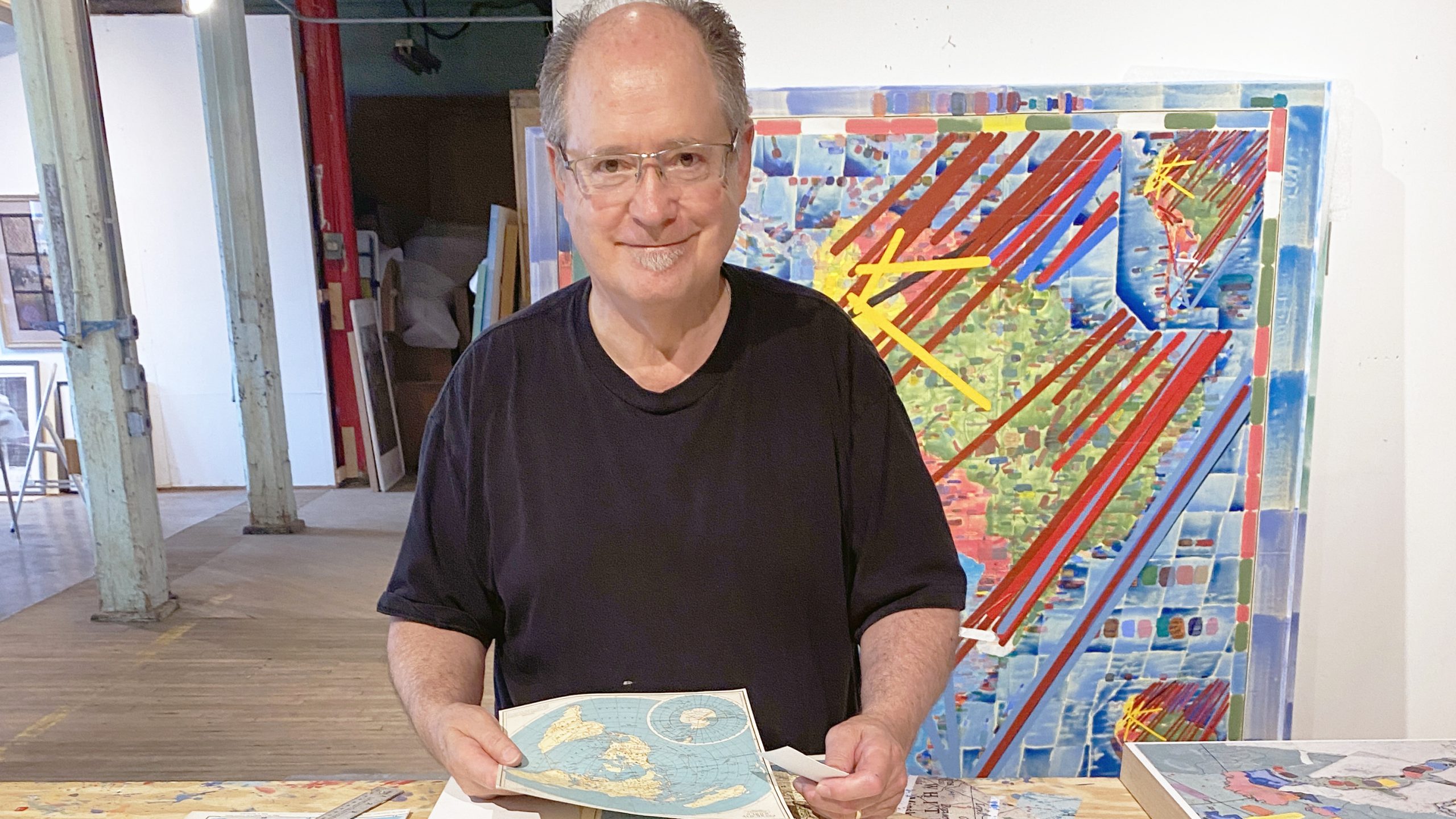 Photograph of artist Dan Mills in his art studio. Dan stands behind a work table with maps in his hands. One of his original paintings is directly behind him and he is pictured in an open building. Dan faces the camera with a smile.