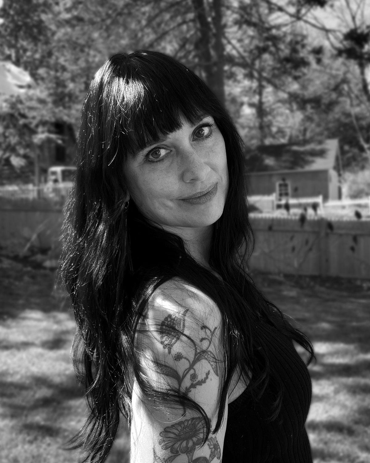 Black and white photograph of artist and author, Summer J. Hart. Summer stands in front of a natural background, facing the camera and looking over her shoulder.