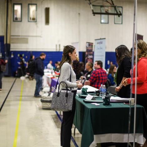 A student stands in front of an employer table in the gym speaking to an employer representative. Other students and employers stand in the background. 