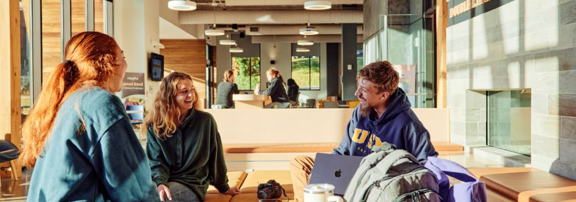 Three students sit in the McGoldrick Center talking to each other with a laptop open.