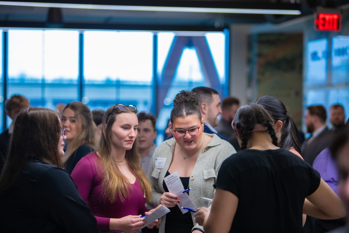 A large group of people gather and talk in front of a window overlooking a city.