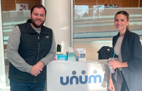 JSP participant and host stand by the front desk at Unum, which has the Unum logo on it.