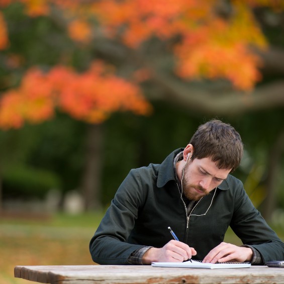 Student writing at picnic table outside with fall foliage in the background