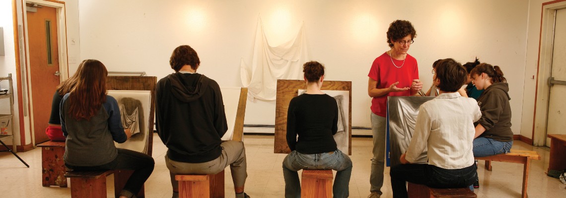 Group of seated students with backs to the camera drawing on easels. Teacher standing in front of them facing camera.