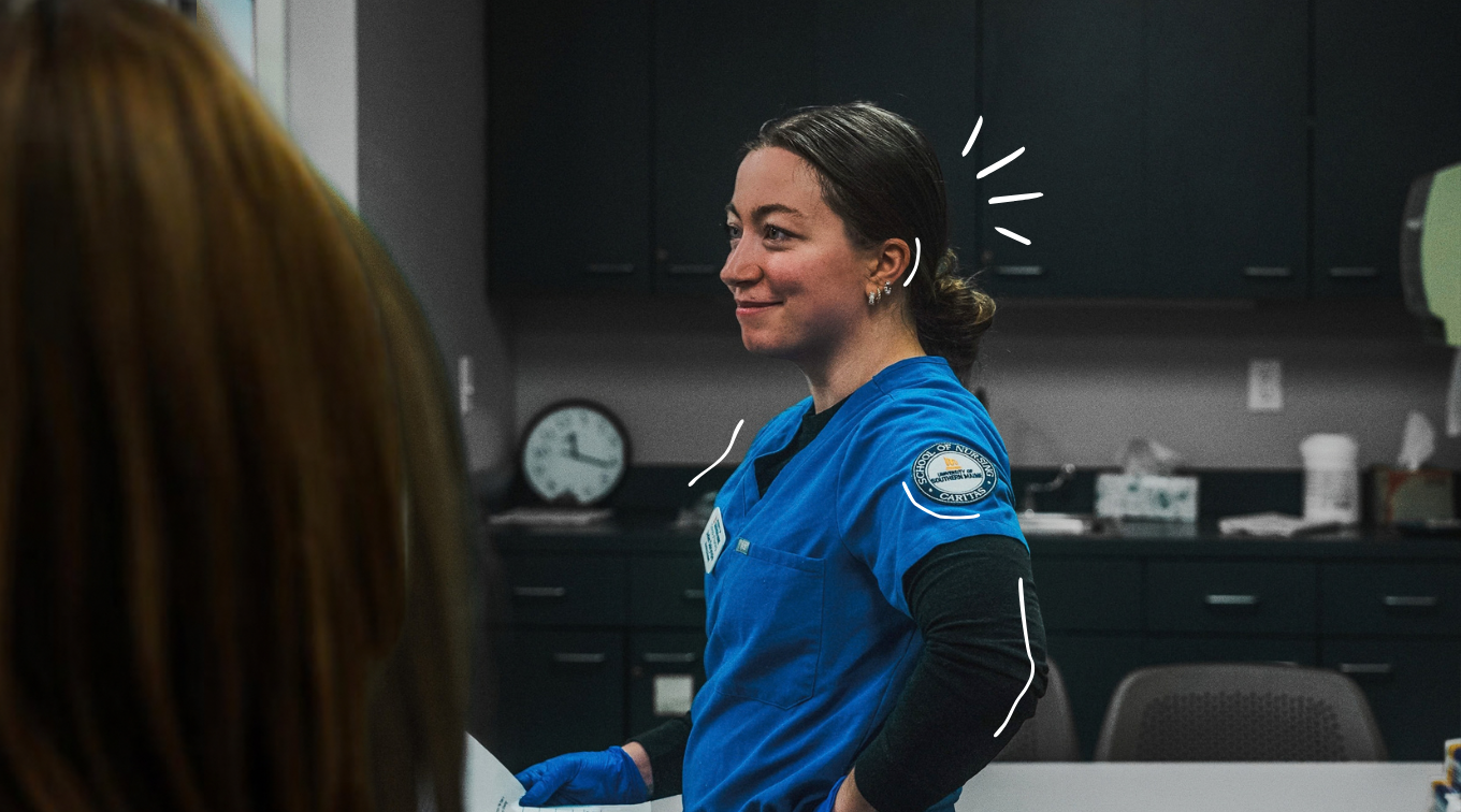 Jaden Dalrymple, a USM nursing graduate in blue School of Nursing scrubs and gloves, smiles and looks off to the side in a nursing lab.