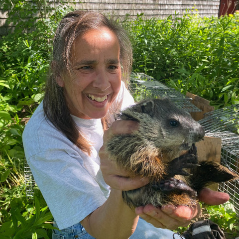 Picture of a woman holding a groundhog in both of her hands with a background of vibrant trees and bushes.