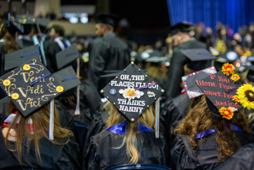 Pictured are three students at the Commencement ceremony with their backs to the camera, showing the viewers their graduation caps which have been decorated with flowers and quotes.