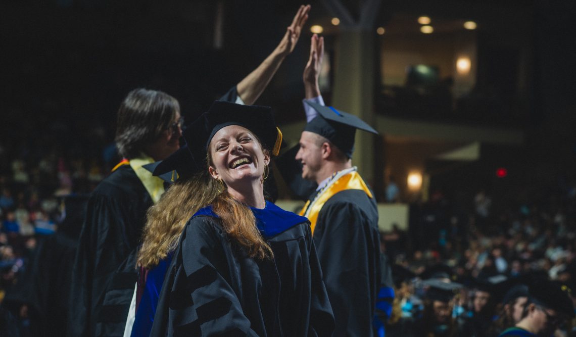 Graduate smiling widely at camera during graduation ceremony. Two people wearing regalia behind the student are high-fiving.